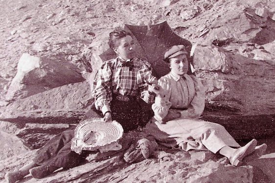 A black and white photograph of two women sitting on rocks in the desert. They are wearing buttoned up shirts and long skirts and are holding a parasol.