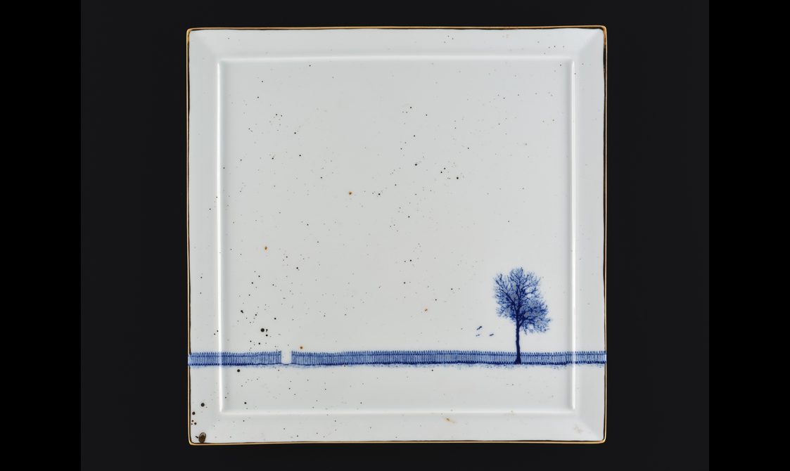 White porcelain square with a blue painting of a fence running horizontally along the square, with a small opening in the fence. A painted blue tree stands in front of a section of the fence.