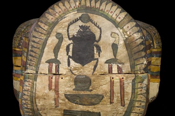 Head end of a wooden coffin, covered in plaster and painted. The decoration on the crown of the head shows a black scarab beetle holding a sun in the centre, surrounded by colourfully painted hieroglyphs.