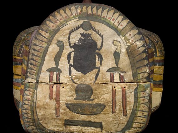 Head end of a wooden coffin, covered in plaster and painted. The decoration on the crown of the head shows a black scarab beetle holding a sun in the centre, surrounded by colourfully painted hieroglyphs.