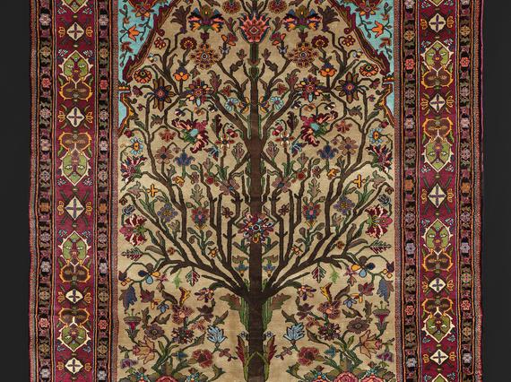 Silk rug depicting an angular tree with branches full of flowers. It is surrounded by a red border with various embellishments.