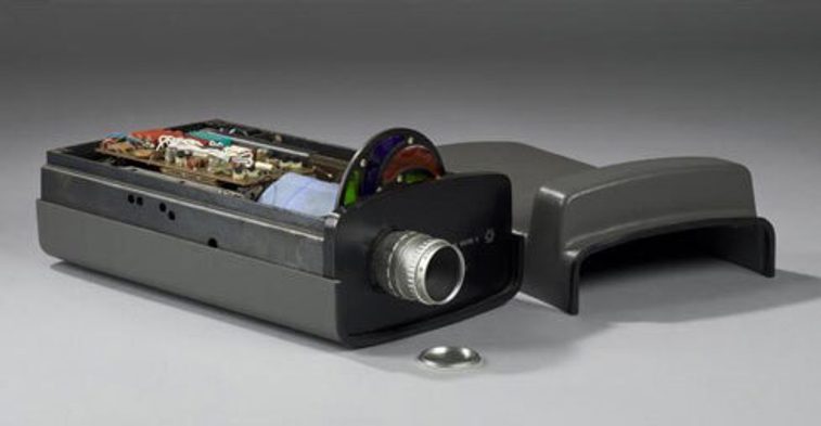 A colour photograph of a piece of rudimentary black camera equipment on its side with the top section exposed. It has a handle and a sense. The exposed area is showing electrical wires and connectors.It's sitting on a grey surface and background.