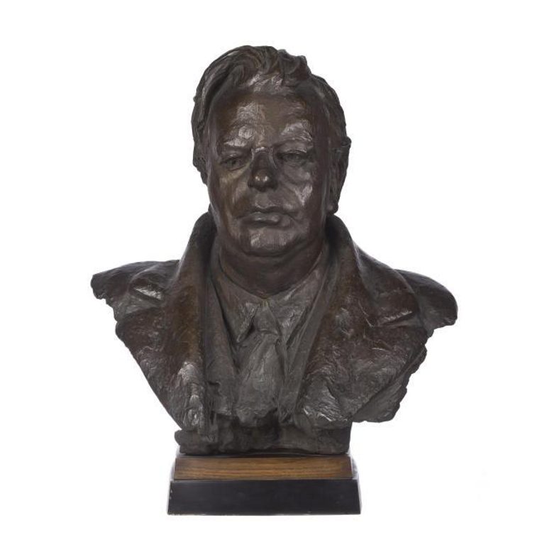 A colour photograph of a dark bronze sculpture bust of a man. It depicts his head and shoulders only. He has a prominent forehead and swept back short hair with a side parting. He's wearing a shirt and toe and a labelled coat.