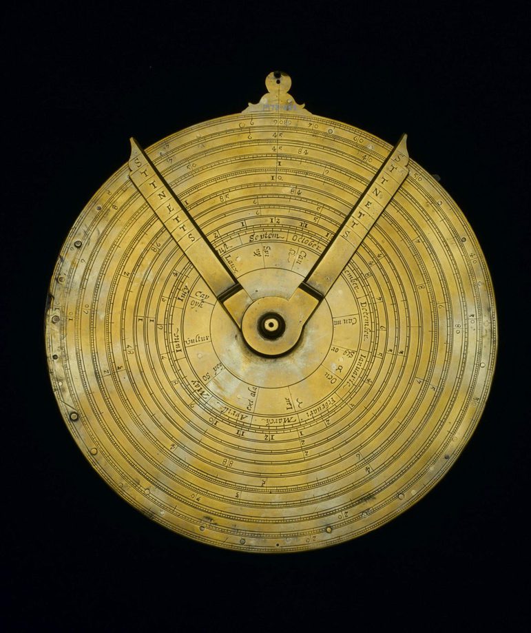 A colour photograph of a yellow metal circular device against a back background. The device has two dials attached at the centre and finishing at the circumference. There are letters engraved on both arms. There are concentric rings on the devoice and engraved measurements.