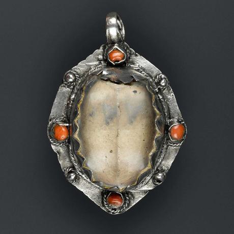An oval silver pendant with a crystal setting. One side of the crystal is damaged. The mount is decorated with four pieces of red coral alternating with four silver balls.