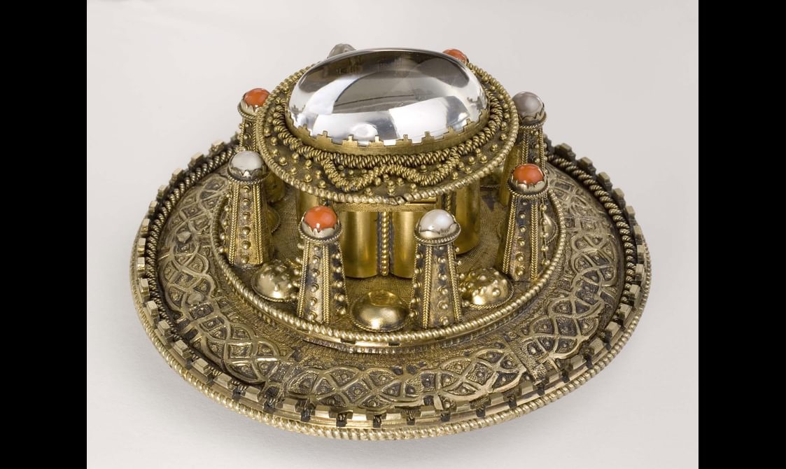A side view of a round gold coloured metal brooch with raised decoration.