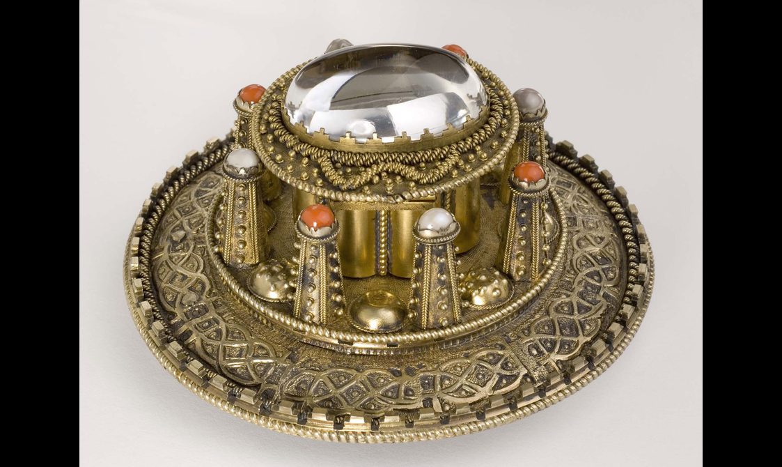 A side view of a round gold coloured metal brooch with raised decoration.