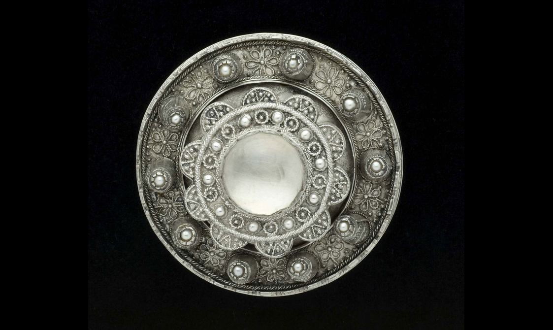 A round silver metal brooch with raised decoration and jewels.
