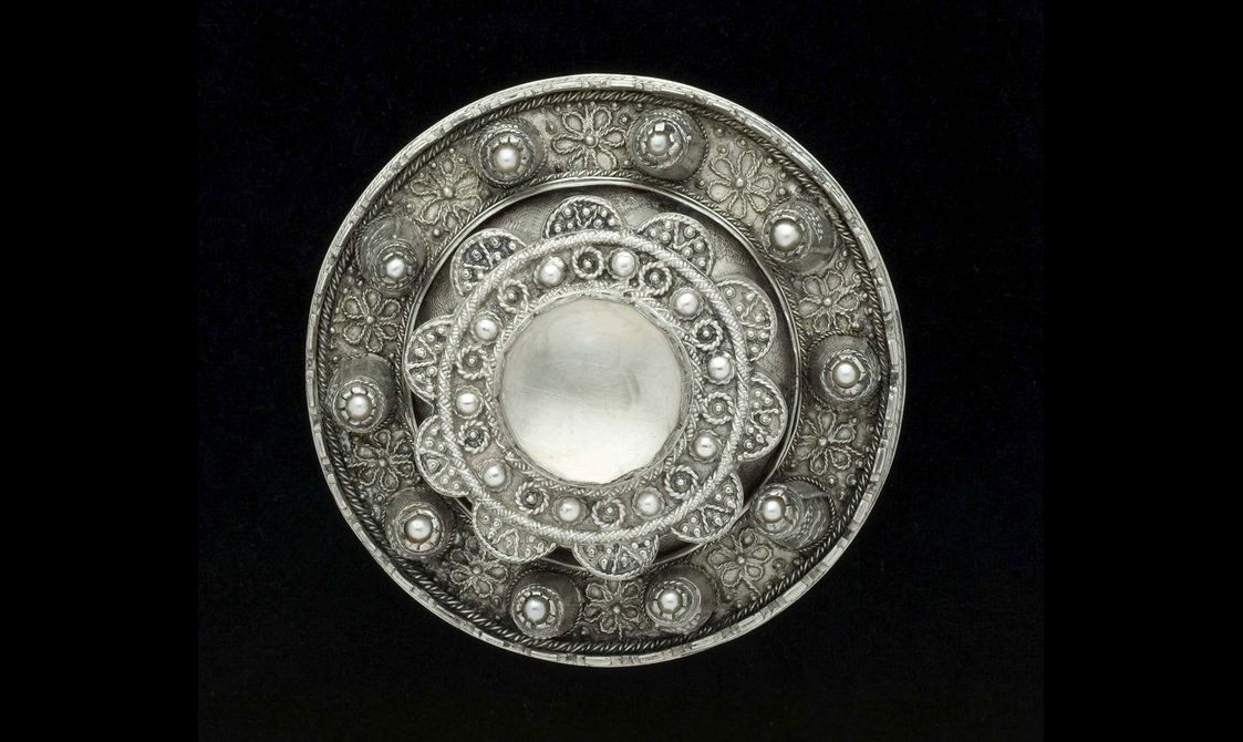 A round silver metal brooch with raised decoration and jewels.