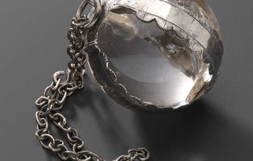 A crystal ball secured in a silver frame with fringed decoration and link chain.