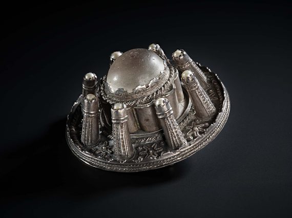 A side view of a round silver metal brooch with raised decoration.