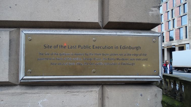 A gold coloured plaque screwed on the end of a brick walled building.