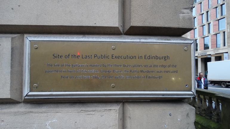 A gold coloured plaque screwed on the end of a brick walled building.