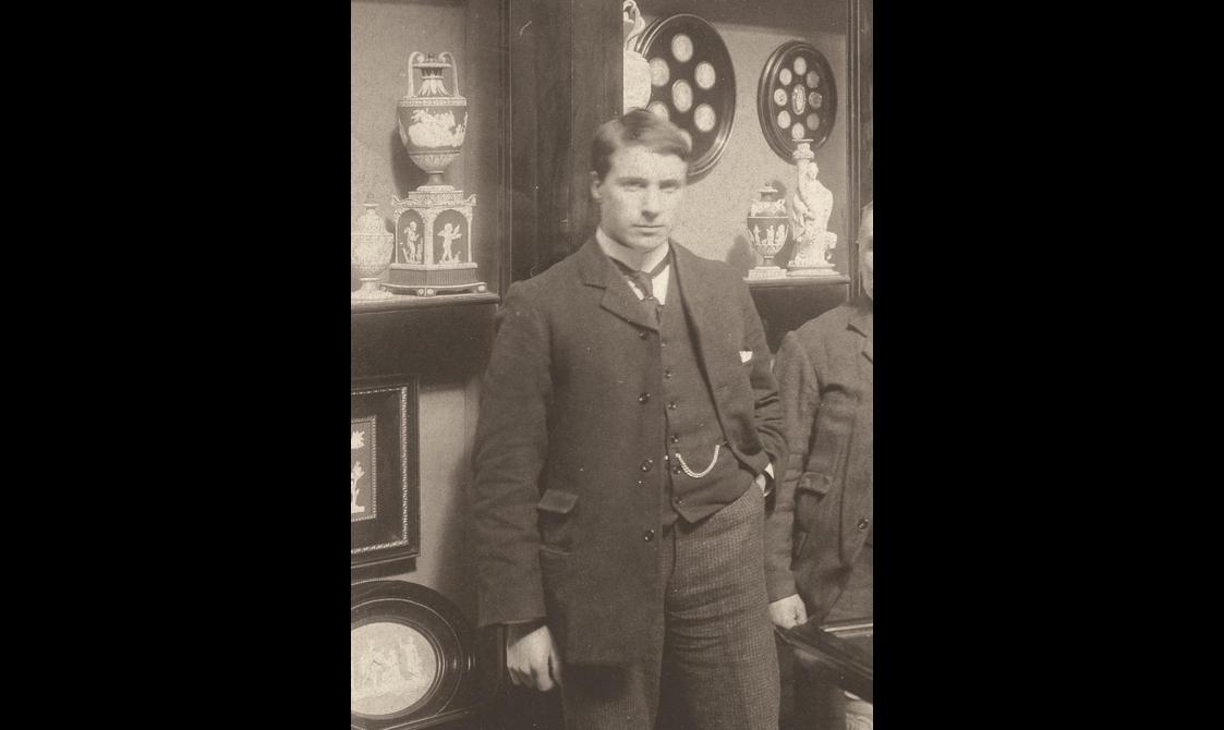 Sepia toned photograph of a man standing in a three-piece suit in front of some shelves of objects.