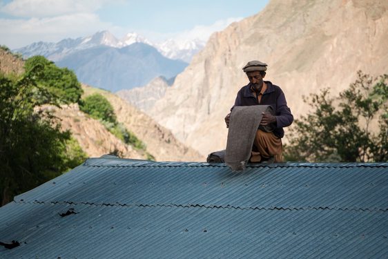 Man sitting on a corrugated metal roof rolling a length of brown fabric. There are large, snowcapped mountains in the background.