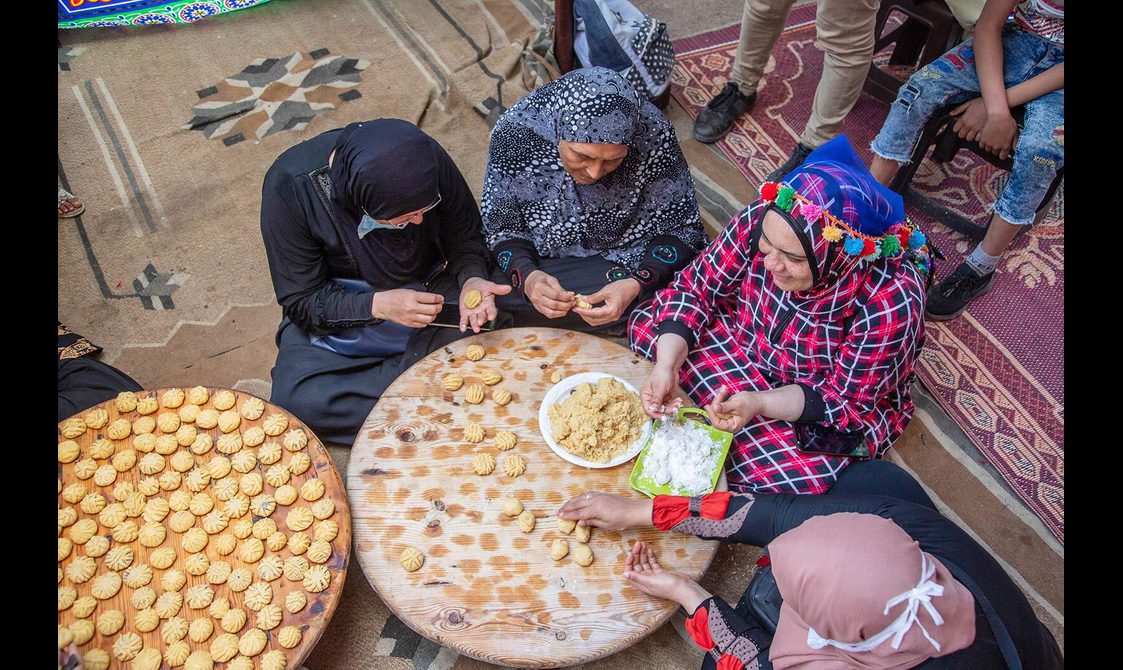 A group of women in traditional hijab dress sitting around a round table making kahk.