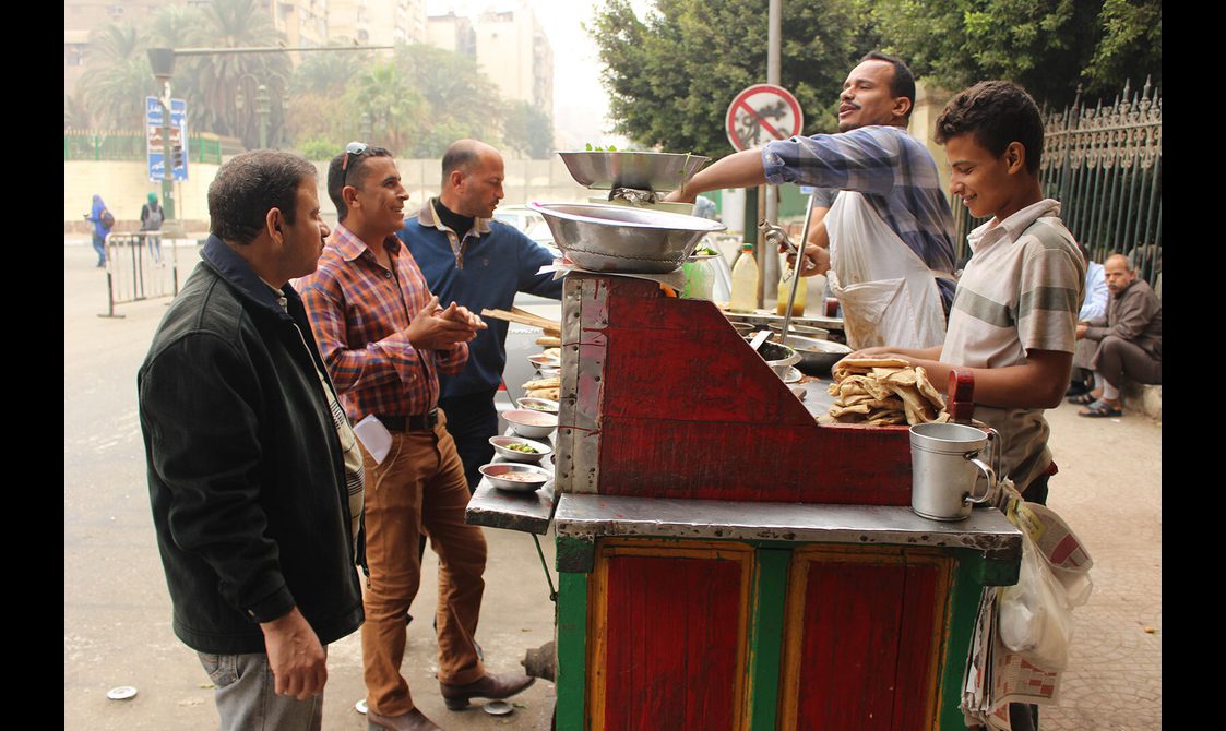 Three people stand at a foul cart while vendors prepare food.