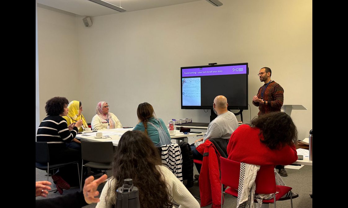 A group of people sit around tables and listen to someone giving a talk about a writing exercise.