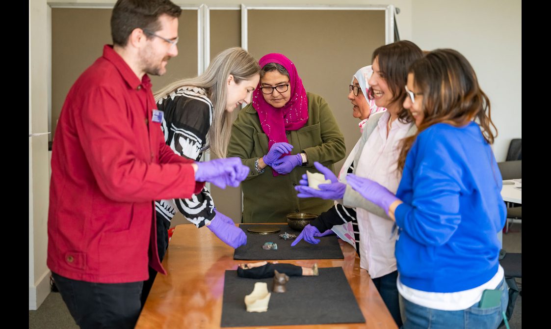 A group of people stand around a table wearing purple nitrile gloves. They are examining different Egyptian objects from the National Collection.