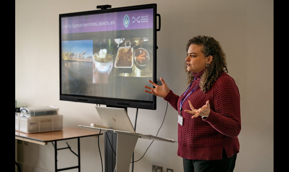 A speaker wearing a red jumper stands next to a screen showing different aspects of Egyptian culture.