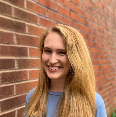A smiling woman dressed in a blue jumper with blonde hair posing against red brick wall.