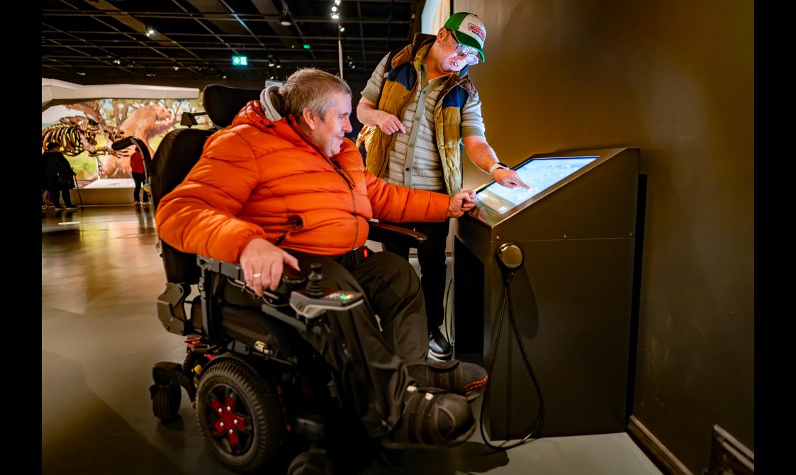 Two people interact with a museum gallery interactive. One person stands wearing a green and white cap. The other sits in a wheelchair and wears an orange jacket. Behind them is a display of a large prehistoric animal skeleton.