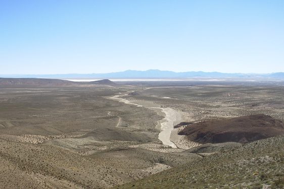 A gently rolling landscape from above covered in scrub bushes with large mountains in the distance.