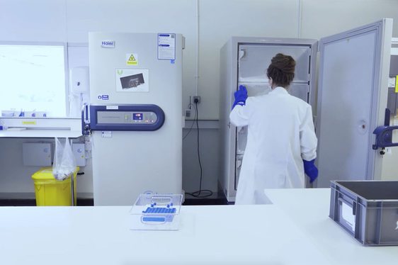 A person in a white lab coat seen from behind opening a large freezer in a mostly white lab room.