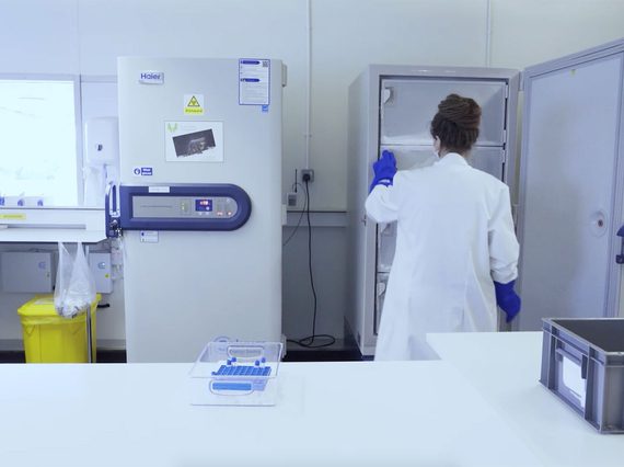 A person in a white lab coat seen from behind opening a large freezer in a mostly white lab room.