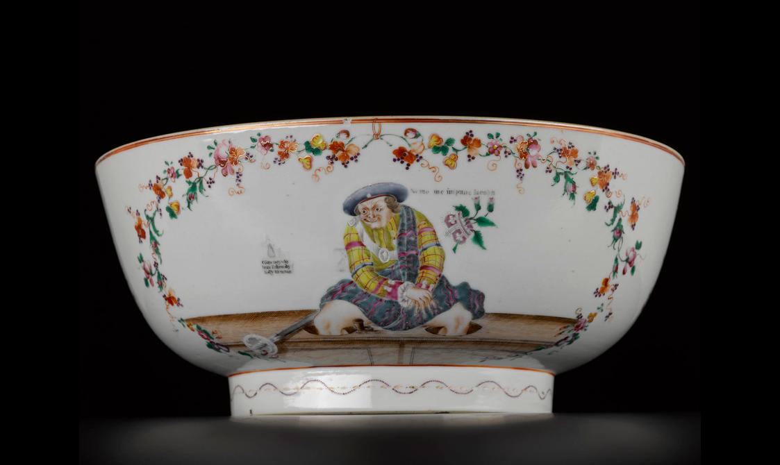 White porcelain bowl shown on a side against black background. Red, yellow and green floral motifs frames a man dressed in yellow, red, green tartan kilt-wear with a blue berret and is seated on a wooden ledge. A crown with floral motifs is suspended to the right hand side of the figure.