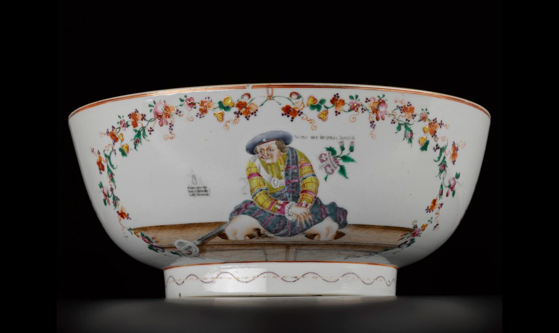 White porcelain bowl shown on a side against black background. Red, yellow and green floral motifs frames a man dressed in yellow, red, green tartan kilt-wear with a blue berret and is seated on a wooden ledge. A crown with floral motifs is suspended to the right hand side of the figure.