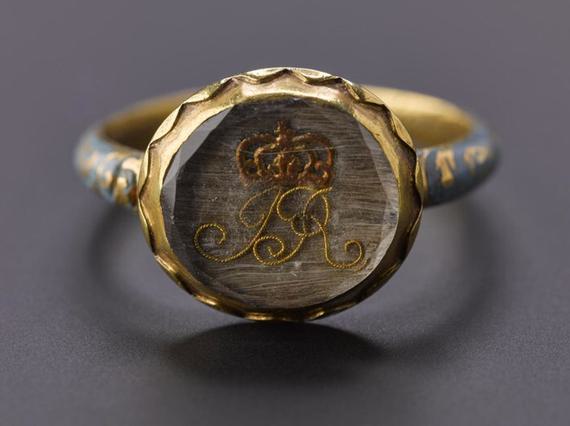 Close up of a circle shaped ring, with a clear gem stone. "JR" initials formed from a light brown thread and a red brown crown design above it.