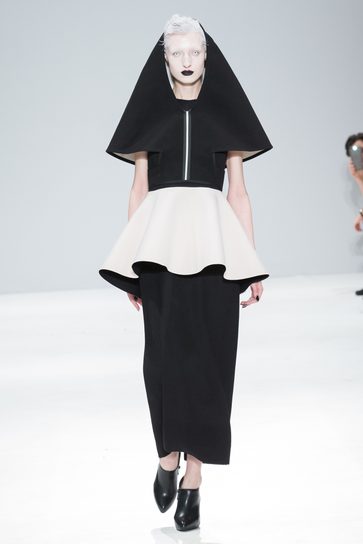 A model with short white hair and black lips wearing a black and white dress, walking down the runway. The dress features a black hood and she is wearing black heeled shoes.