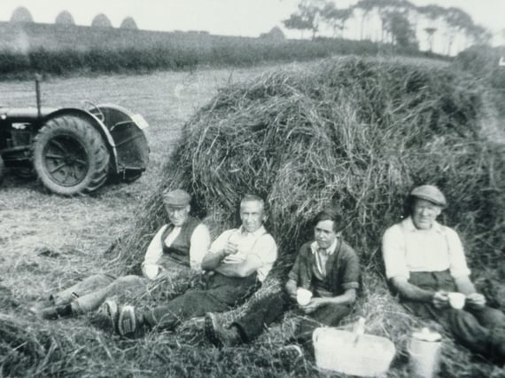 A black and white photograph of four farmers sitting against a haybale with cups in their hands on a farm.