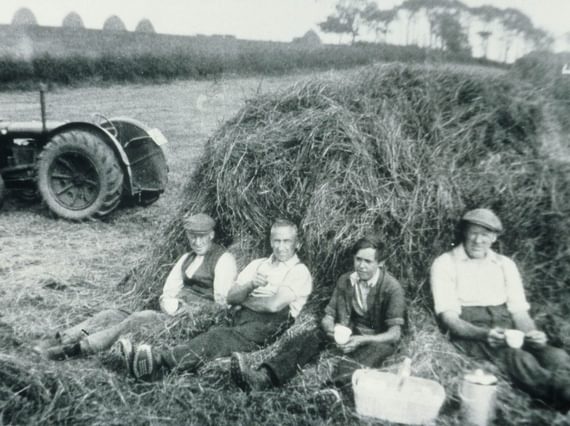 A black and white photograph of four farmers sitting against a haybale with cups in their hands on a farm.