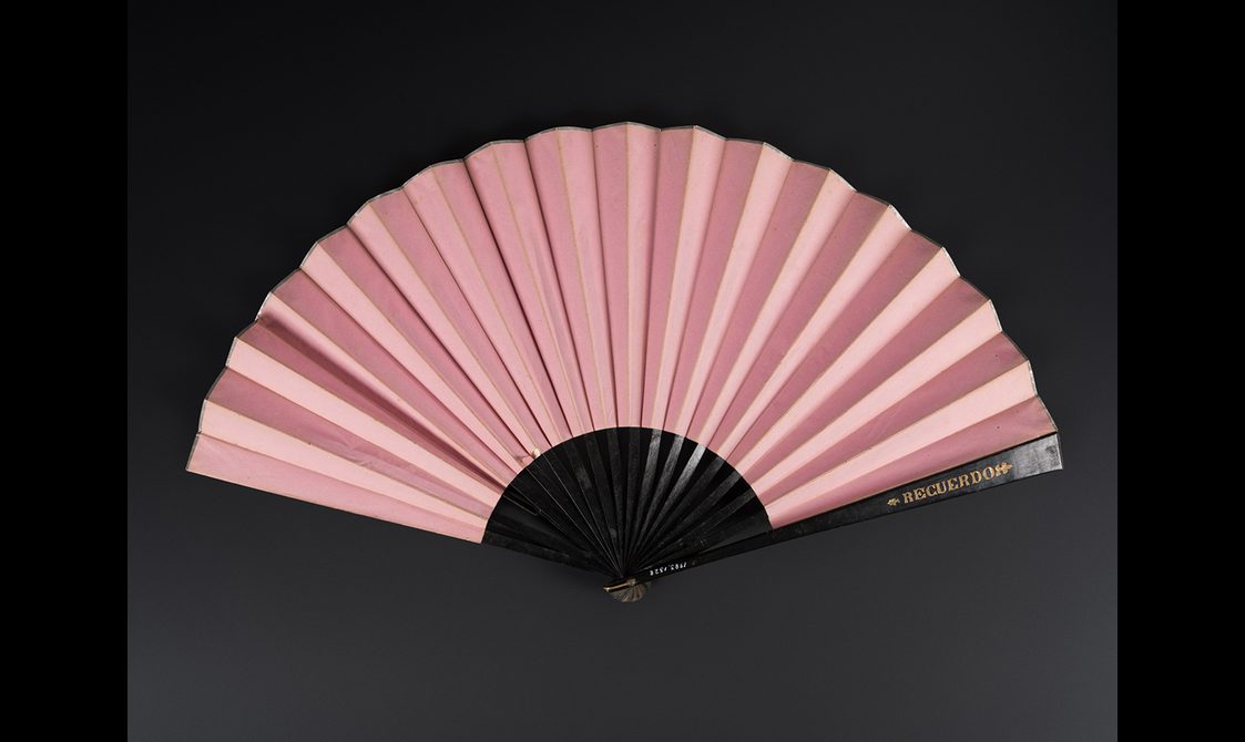 Fan with wooden sticks and guards painted black with gilt detailing. The body of the fan is plain pink paper. The guard has the inscription 'RECUERDO'.