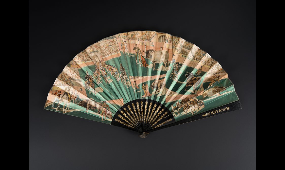 Fan with wooden sticks and guards painted black with gilt detailing. One guard with inscription 'ESPANA'. Fan leaf in metallic paper printed with a bullfighting scene