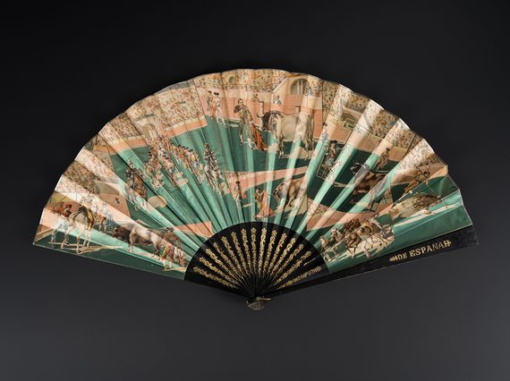 Fan with wooden sticks and guards painted black with gilt detailing. One guard with inscription 'ESPANA'. Fan leaf in metallic paper printed with a bullfighting scene