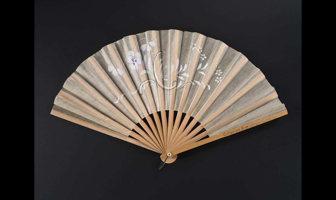 Fan, of paper with wooden sticks and guards. printed in gold with 'Criterion', leaf painted with a purple and floral spray and with glitter highlight swirls.