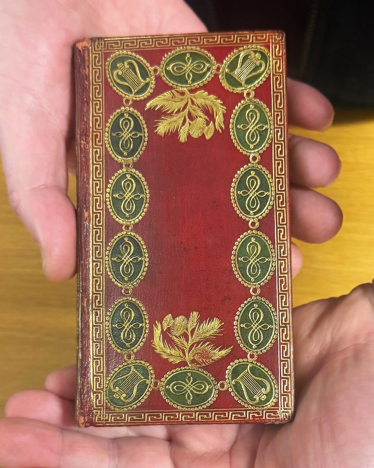 Hands holding a small, red covered book, with details in red, black and gold around the edge of the cover. There's a wooden floor in the background.