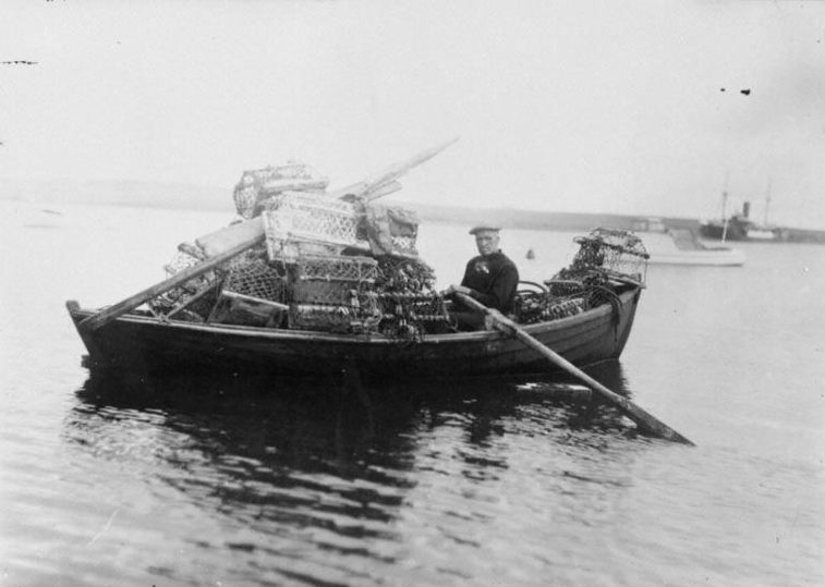 Person sitting in a wooden boat filled with lobster traps on calm water, with another boat visible in the background.