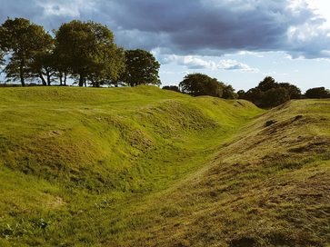 Looking down a deep ditch cutting across a green open field on a cloudy day, with earthen ramparts on either side.
