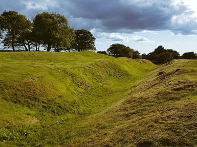 Looking down a deep ditch cutting across a green open field on a cloudy day, with earthen ramparts on either side.