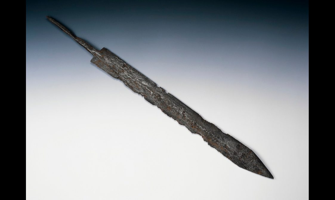 A classic Roman gladius with triangular point, straight blade and narrow tang set against a grey-blue background.