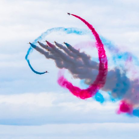 Nine planes fly in formation with blue and red smoke trailing making patterns in the sky.