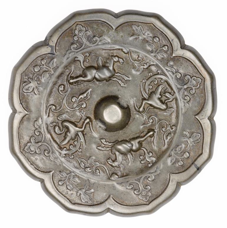 A silver round object with a petal shape around the edge. It has horse and floral patterns on its surface.