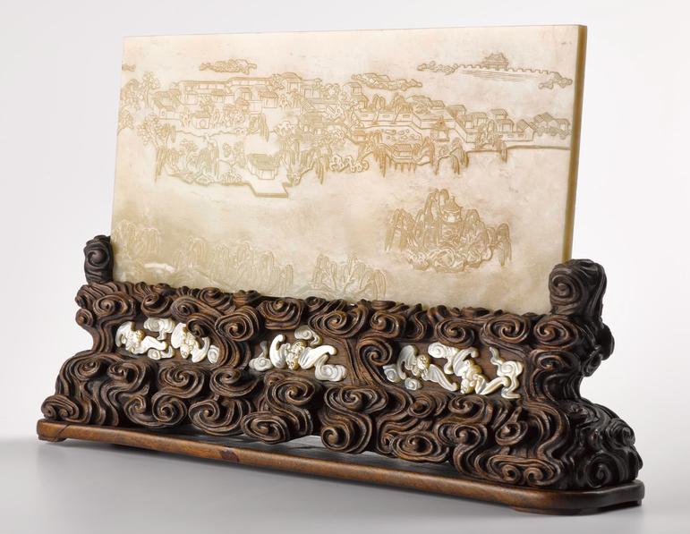 An elaborately carved long wooden stand holding a rectangular piece of pal green jade engraved with a landscape scene.