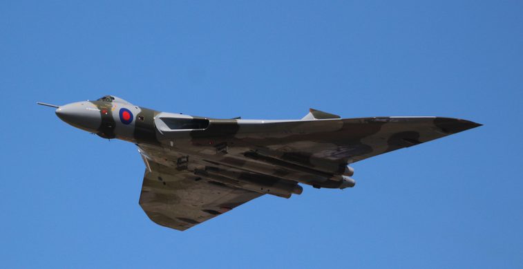An aircraft with light grey and khaki green pattern on its surface flying in a blue sky.