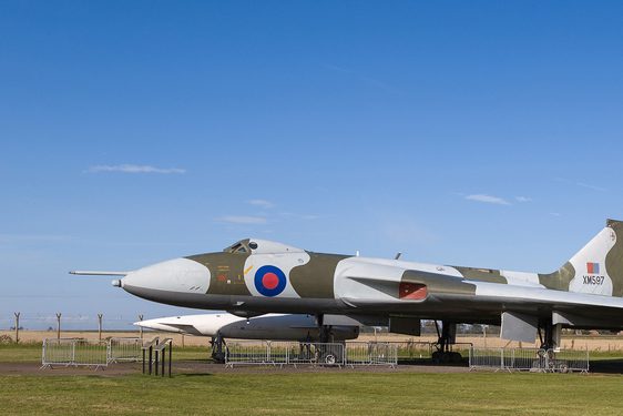 An aircraft with light grey and khaki green pattern over its surface and a blue and red round target near its nose. It sits on the ground surrounded by fencing in the countryside.