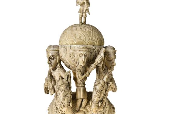 Tall off-yellow ivory sculpture. Two figures on horseback flack two orbs, on atop the other. A small human figure stand between the orbs, another crowns the sculpture.